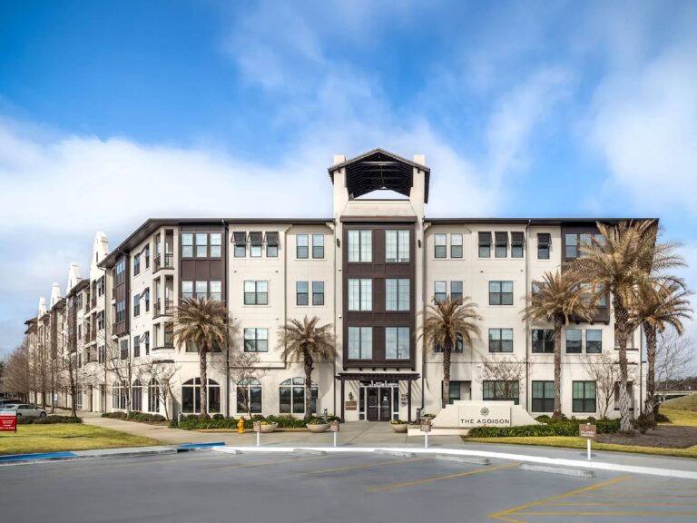 Exterior view of The Addison apartment community in Baton Rouge featuring contemporary architecture, landscaped grounds, palm trees, and a grand residential entrance.