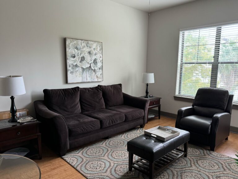 Comfortable living room at The Addison featuring a plush sofa, leather accent chair, coffee table, decorative artwork, and large window with natural light.