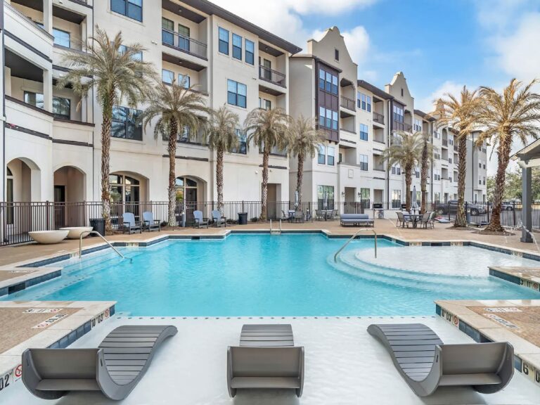 Resort style swimming pool at The Addison featuring in-water lounge chairs, palm trees, sun deck seating, and luxury apartment buildings surrounding the courtyard.