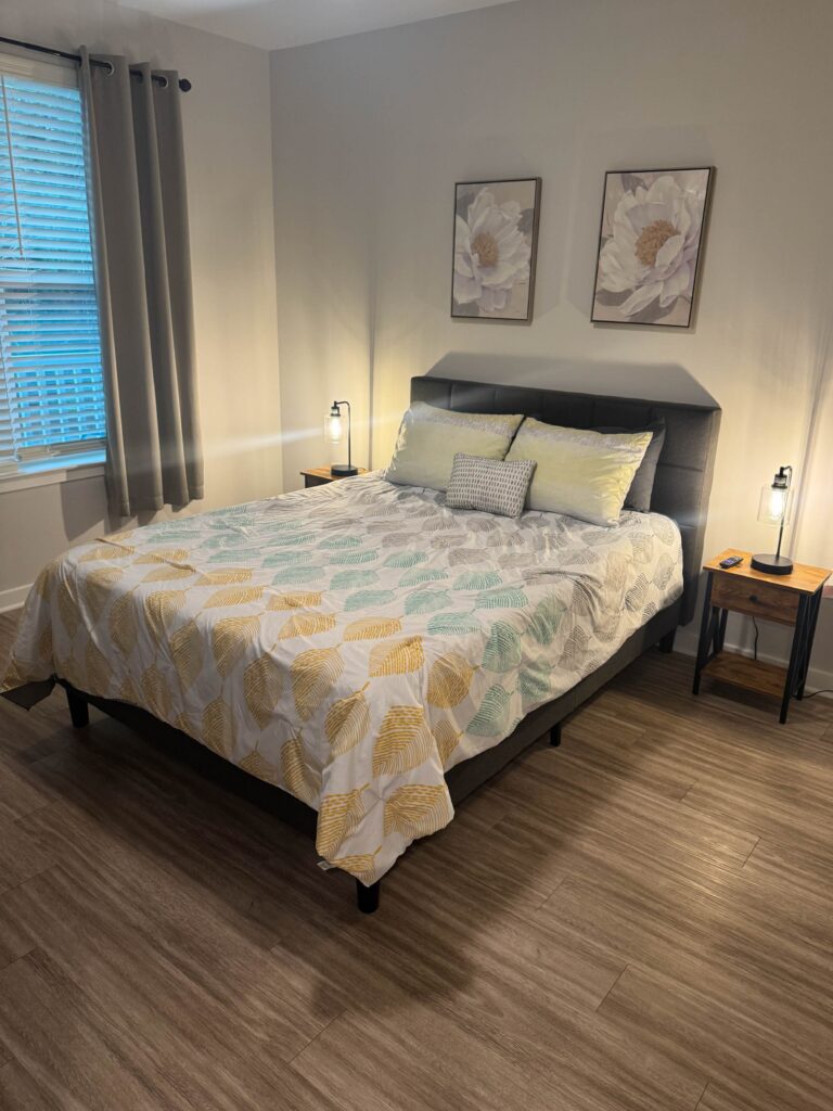 Spacious primary bedroom featuring a king-size bed, contemporary furnishings, warm accent lighting, large window with blackout curtains, and luxury vinyl plank flooring for a comfortable and relaxing retreat.