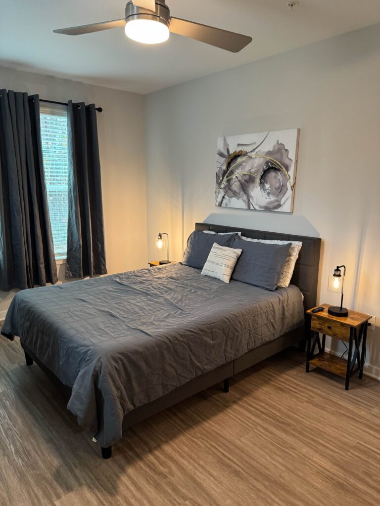 Contemporary bedroom featuring a king-size bed with modern gray bedding, bedside tables with accent lighting, ceiling fan, blackout curtains, and stylish abstract wall art, creating a comfortable and relaxing retreat.