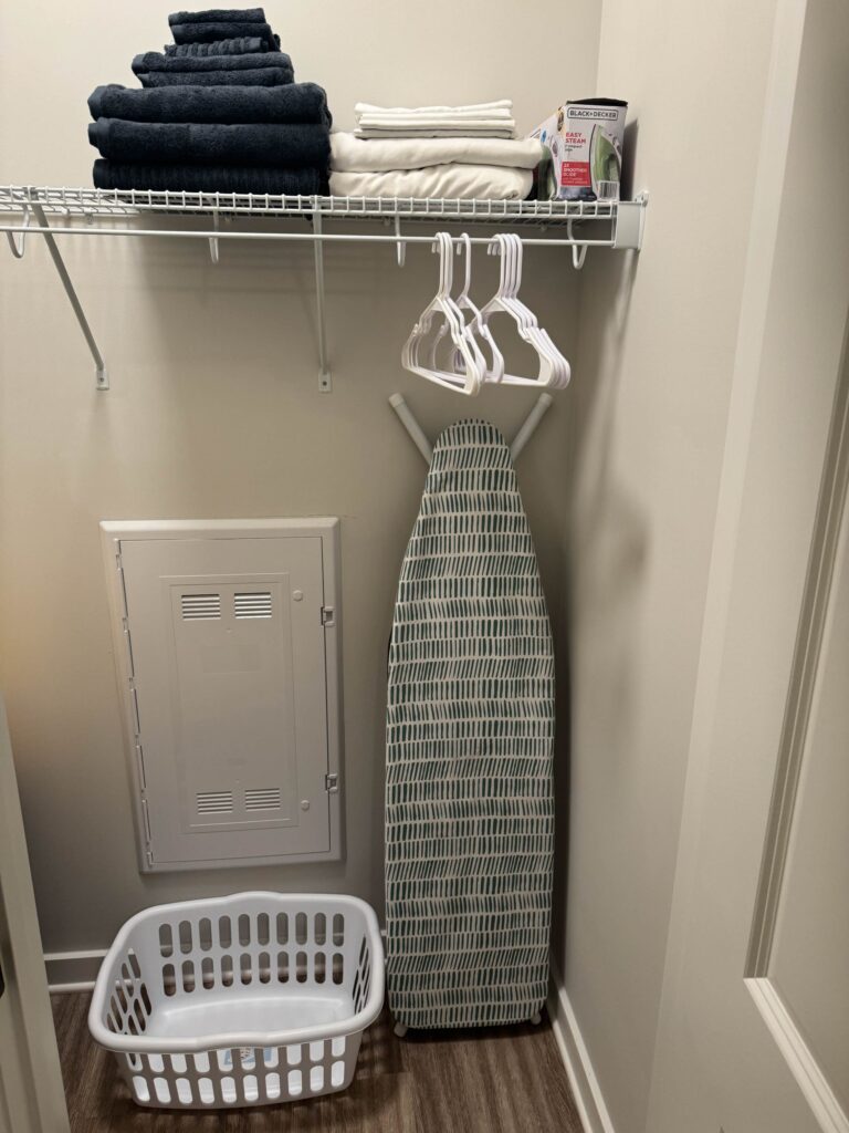 Walk-in closet featuring hanging space, storage shelving, an ironing board, laundry basket, hangers, fresh linens, and additional household essentials for extended stay convenience.