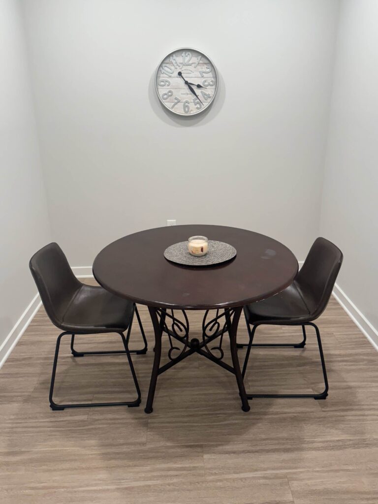 Dedicated dining area featuring a round dark wood table with seating for two, contemporary upholstered chairs, decorative centerpiece, and a large wall clock, creating a comfortable space for meals, work, or conversation.