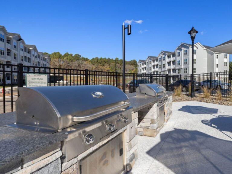 Outdoor grilling pavilion at Alexander Crossing featuring stainless steel gas grills, stone countertops, ample preparation space, and a landscaped community gathering area surrounded by modern apartment buildings.