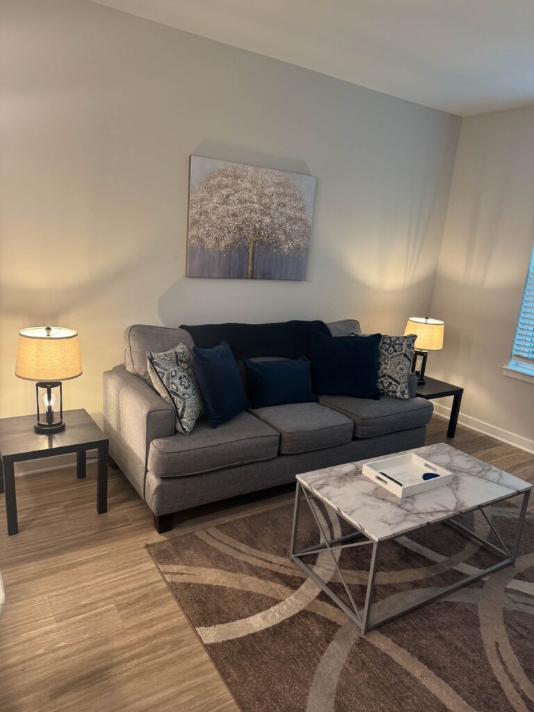 Modern living room at Alexander Crossing featuring a comfortable gray sofa, decorative accent pillows, marble style coffee table, contemporary lighting, and a warm, inviting atmosphere designed for relaxation and everyday living.