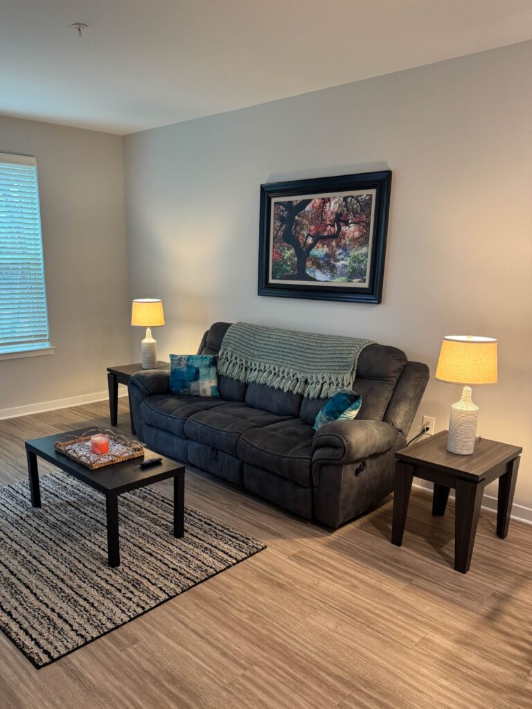 Welcoming living room at Alexander Crossing featuring a plush reclining sofa, decorative accent pillows, soft lighting, framed artwork, and a comfortable seating area designed for relaxation and everyday living.