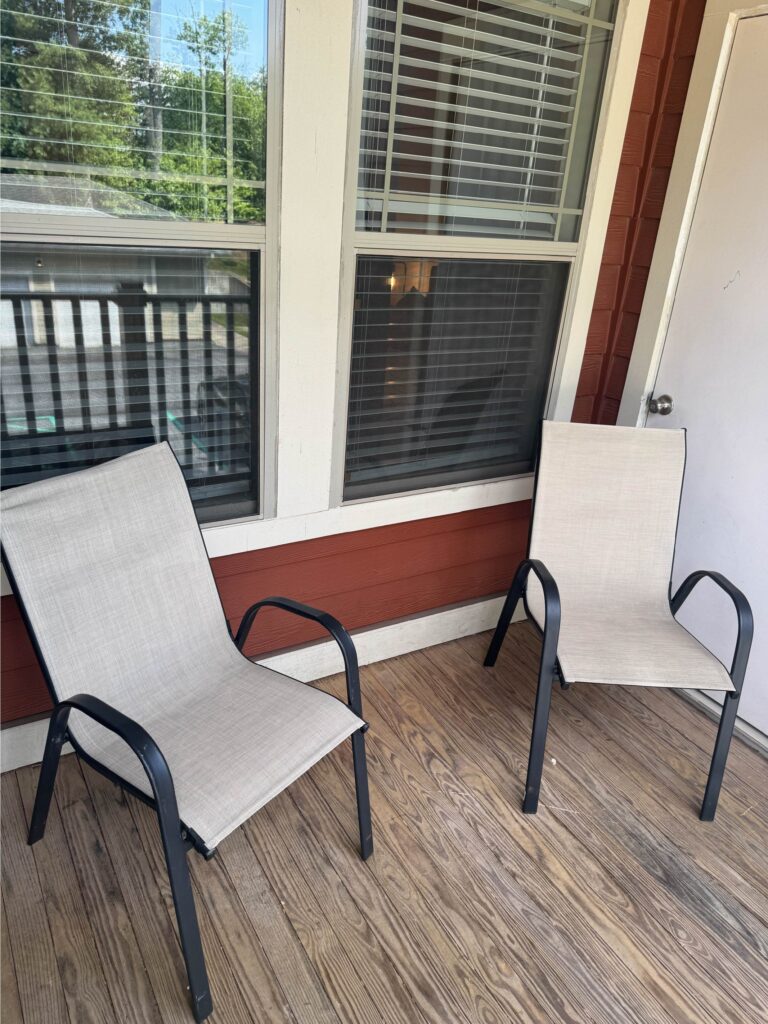Private covered balcony featuring two outdoor chairs, wood plank flooring, and views of surrounding trees and the community grounds.
