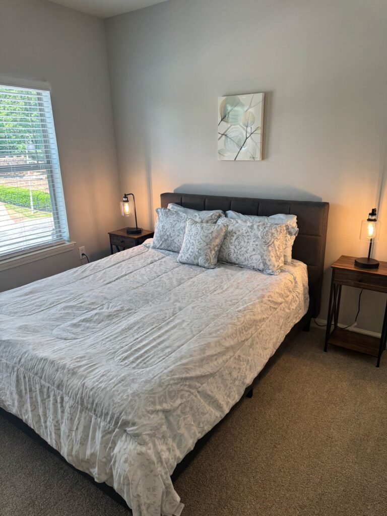 Primary bedroom featuring a queen-size bed with patterned bedding, matching nightstands, decorative lighting, wall art, and a large window providing natural light.
