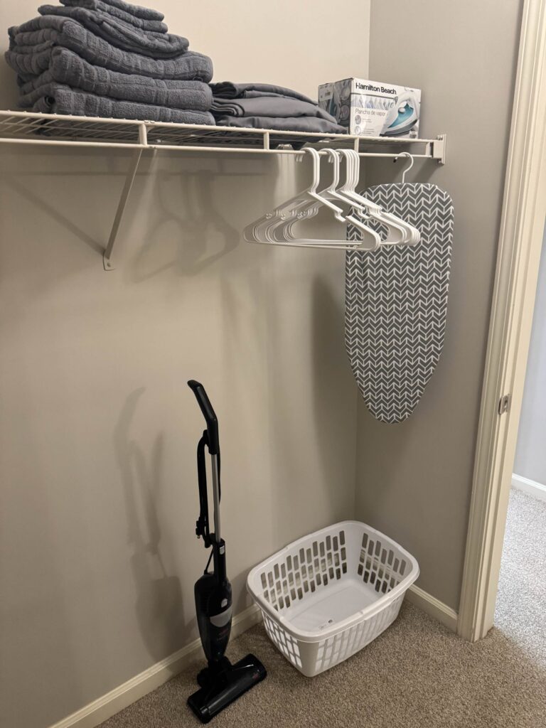 Walk-in closet featuring shelving with extra linens, hanging space with hangers, ironing board, laundry basket, vacuum, and household essentials.