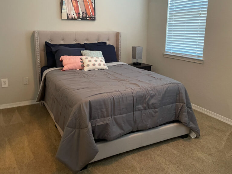 Bedroom at Ballantrae Apartments with upholstered bed, gray bedding, accent pillows, carpet flooring, and furnished apartment setup
