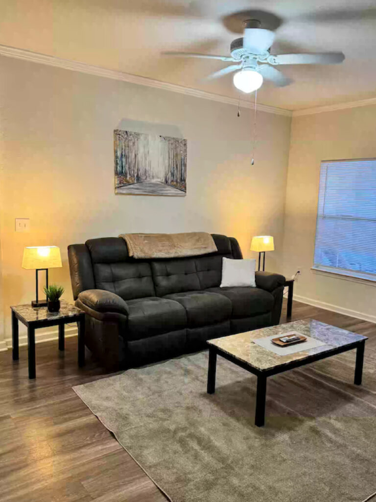 Living room at Ballantrae Apartments with reclining sofa, coffee table, accent lighting, ceiling fan, and furnished extended stay setup