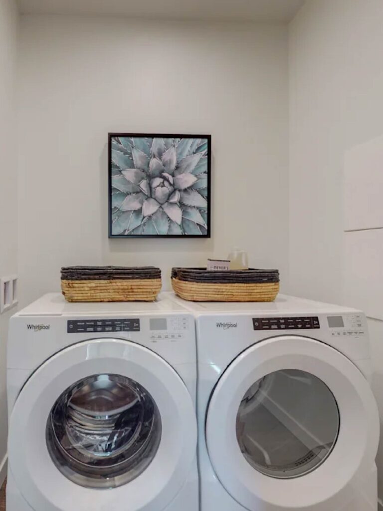 In-unit laundry area at Cielo Luxury Living featuring a full-size Whirlpool front-loading washer and dryer with decorative storage baskets and modern wall art.