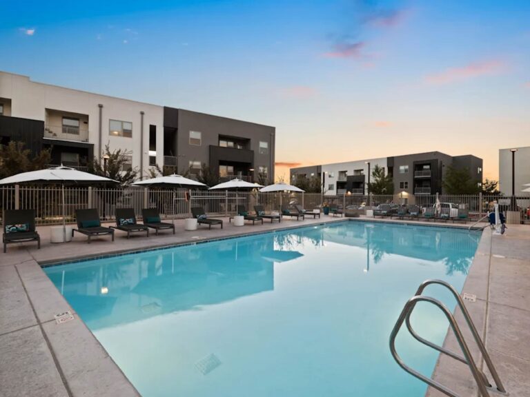 Resort-style outdoor swimming pool at Cielo Luxury Living featuring expansive deck space, lounge seating, sun umbrellas, and modern apartment buildings surrounding the pool area at sunset.