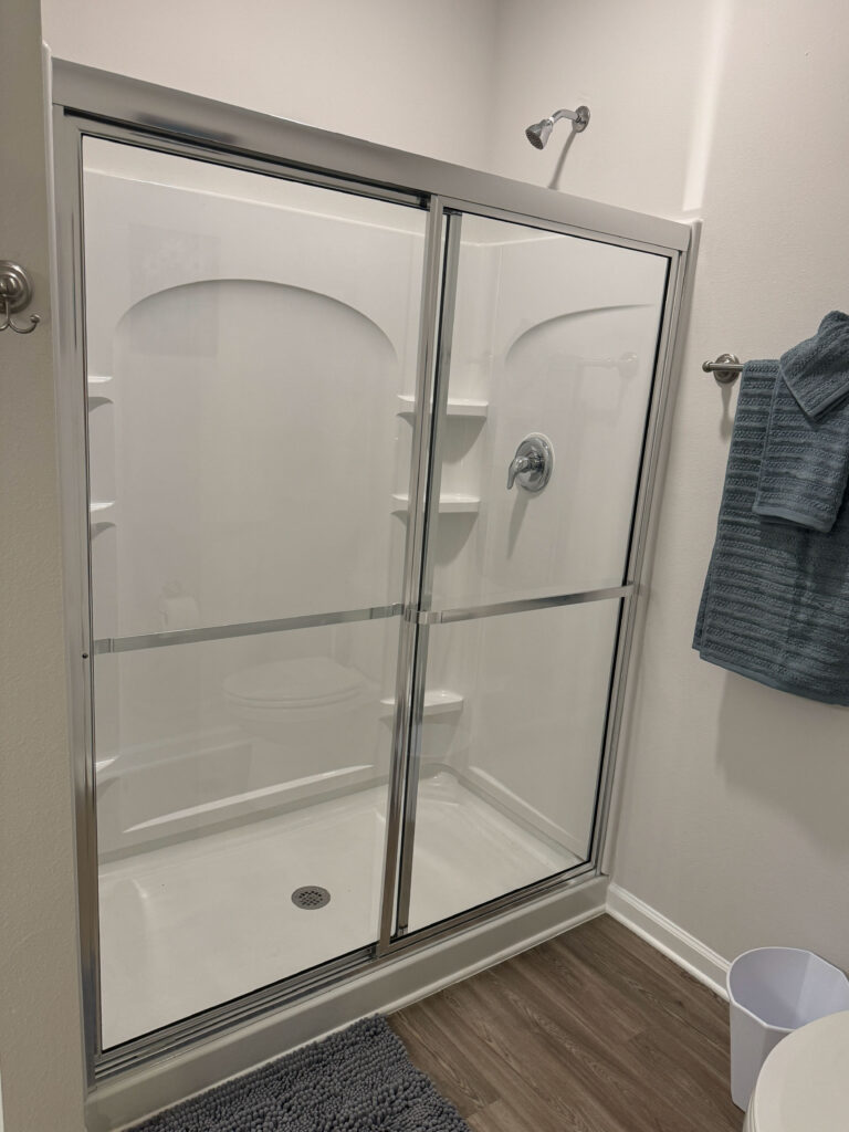 Modern bathroom shower with sliding glass doors, built-in shelving, and wood-style flooring at Covey Homes.