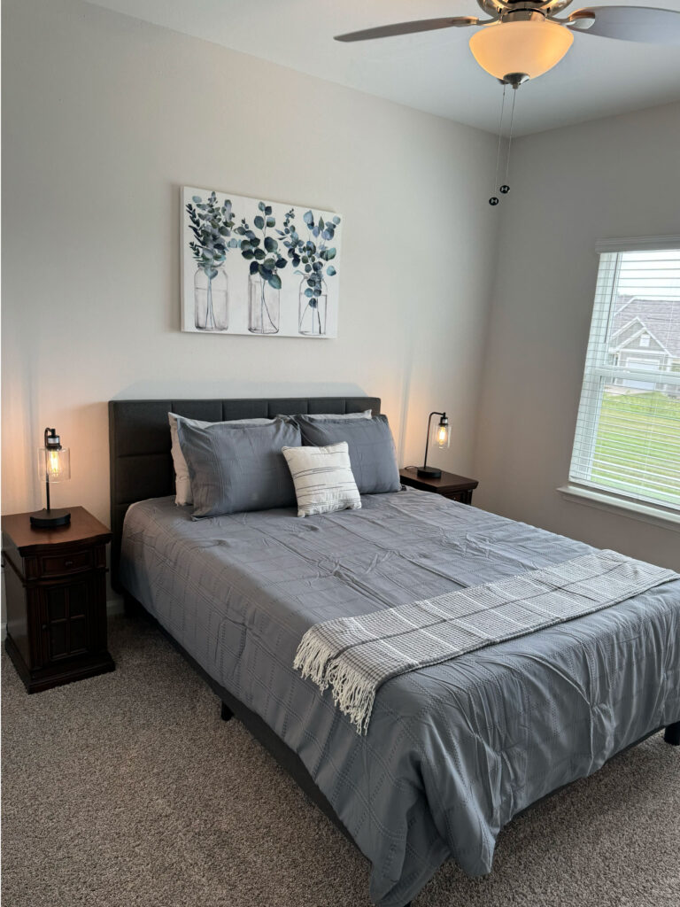 Modern bedroom at Covey Homes featuring gray bedding, bedside lighting, ceiling fan, and large window with natural light.