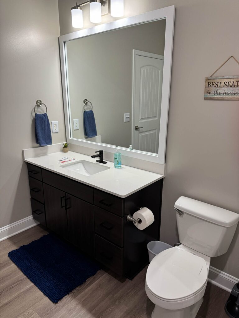 Modern bathroom at Creekstone at RTP featuring a spacious vanity with quartz countertop, large framed mirror, contemporary lighting, dark wood cabinetry, and updated fixtures.