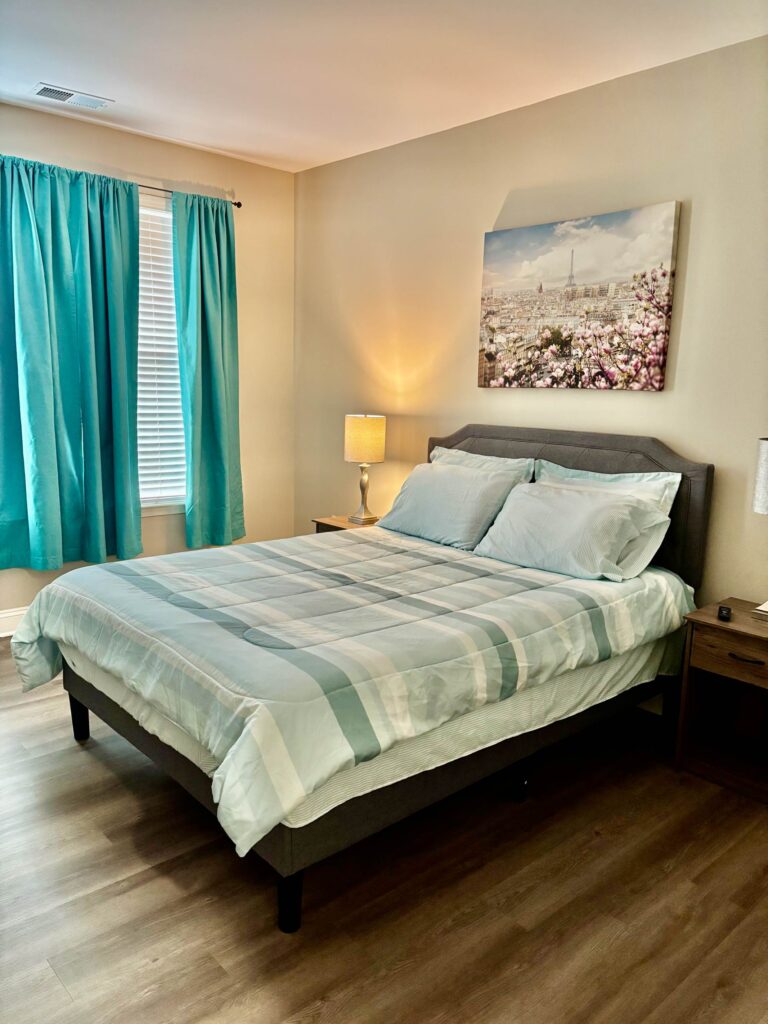 Primary bedroom at Creekstone at RTP featuring a queen size bed, hardwood style flooring, bedside tables with lamps, large window, and contemporary décor with vibrant accent curtains.