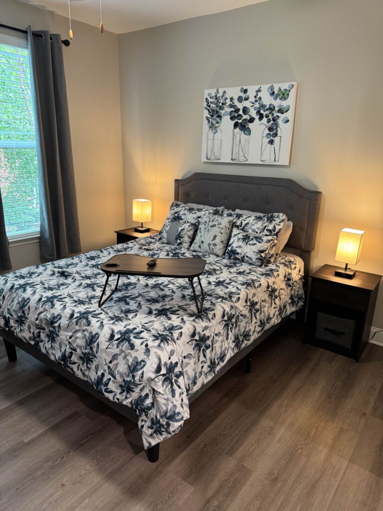 Spacious bedroom at Creekstone at RTP featuring a king size bed with floral bedding, matching nightstands, bedside lamps, hardwood style flooring, and large windows providing natural light.