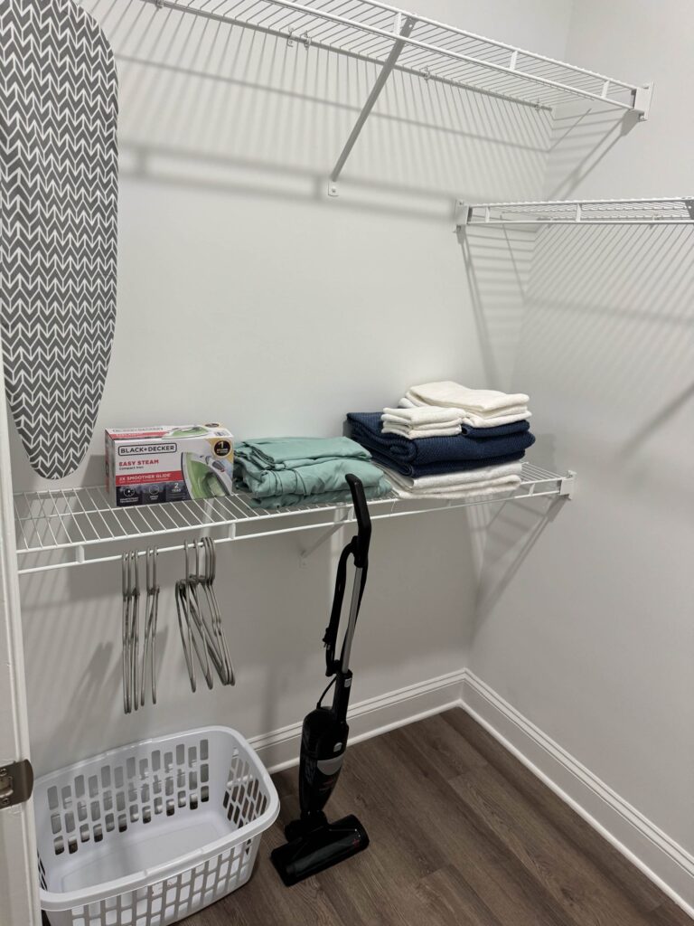 Walk in closet at Creekstone at RTP featuring wire shelving, hanging space, iron and ironing board, extra linens, laundry basket, and a vacuum cleaner for guest convenience.