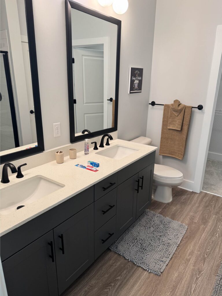 Spacious primary bathroom featuring a double-sink vanity with quartz countertops, dark gray cabinetry, black fixtures, large framed mirrors, and ample storage.