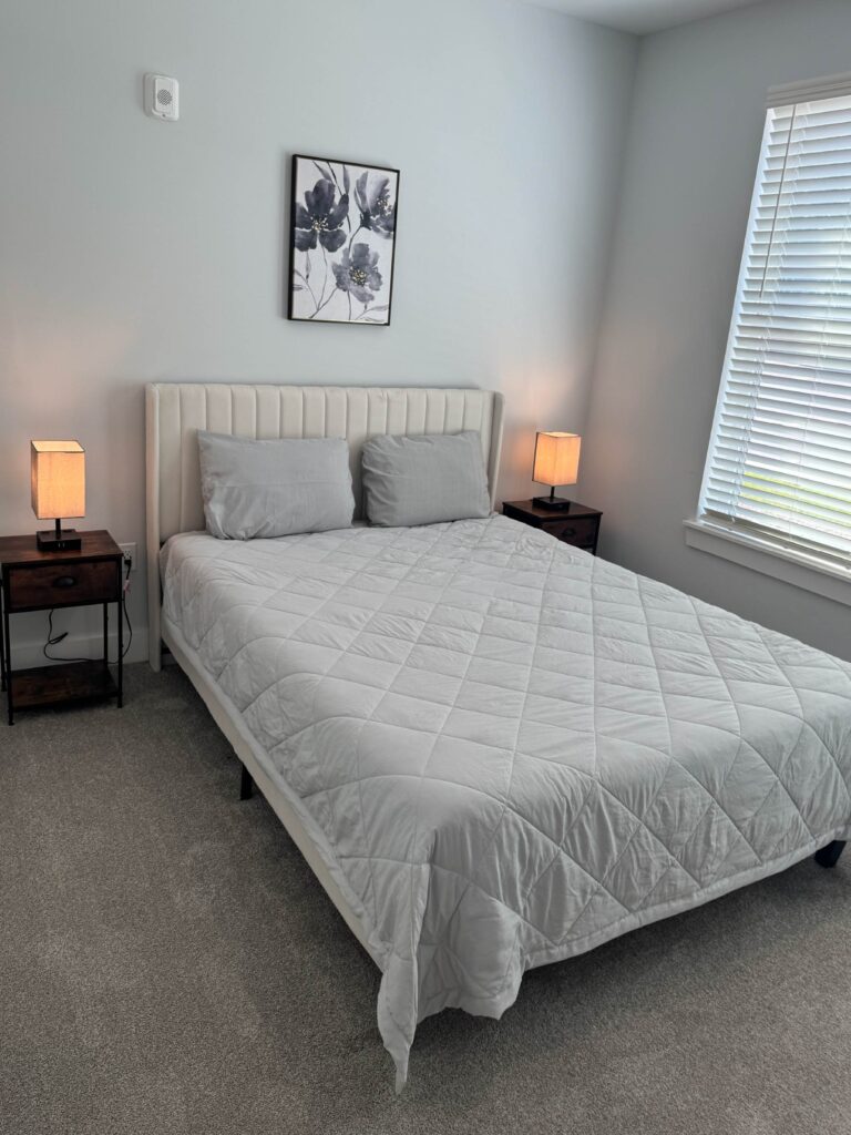 Bright and modern bedroom featuring a queen-size bed with a white upholstered headboard, gray bedding, matching nightstands with lamps, soft carpeting, and a large window with natural light.