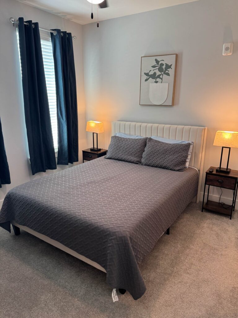 This thoughtfully furnished bedroom at Ellison at the Preserve offers a relaxing retreat after a busy day in Asheville. The room features a comfortable queen-size bed, soft carpeting, bedside lighting, blackout curtains, and tasteful décor that creates a warm and inviting atmosphere. Ample natural light fills the space during the day, while the calming color palette promotes rest and relaxation in the evening. Whether staying for corporate travel, relocation, healthcare assignments, government deployment, or a long-term project, guests can enjoy a private, comfortable sleeping environment designed to support both productivity and well-being throughout their stay.