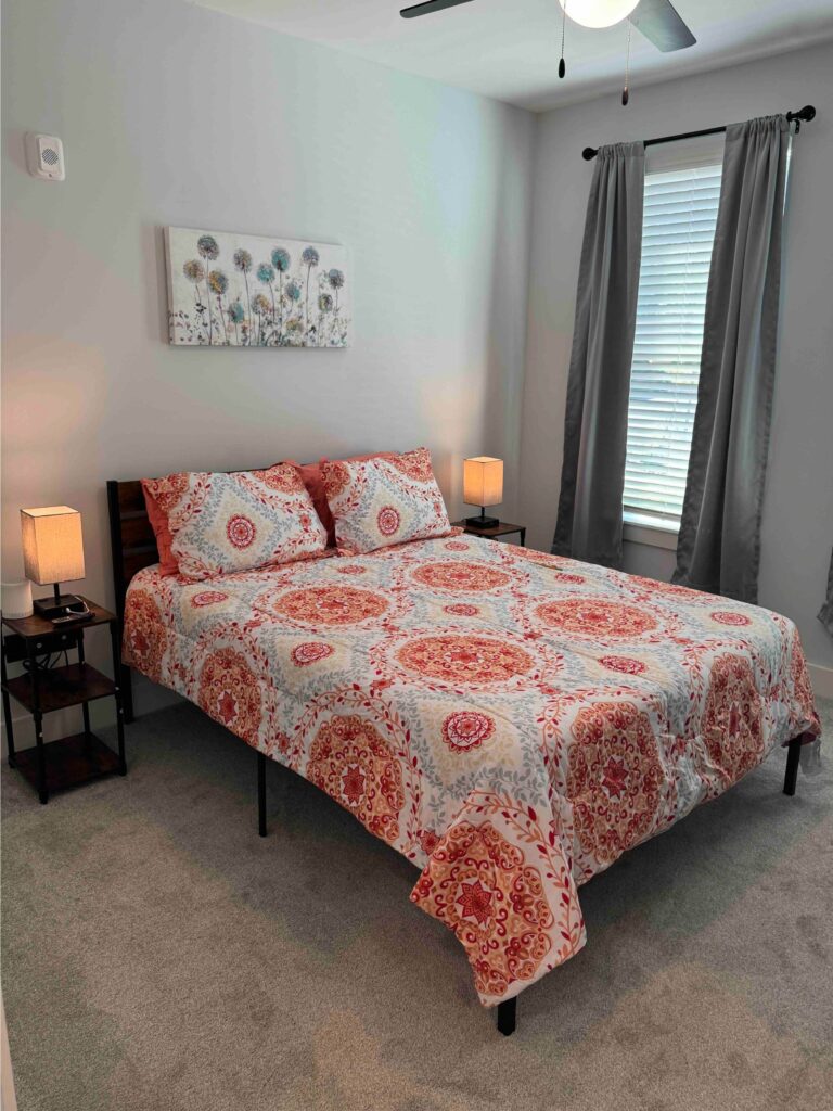 Comfortable bedroom featuring a queen-size bed with patterned bedding, bedside tables with lamps, blackout curtains, soft carpeting, and contemporary wall art.