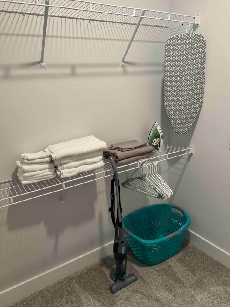 Spacious walk-in closet featuring wire shelving, hanging space, fresh linens, hangers, ironing board, iron, laundry basket, and a cordless vacuum.
