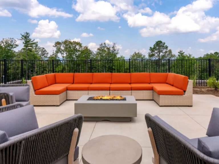 Outdoor community lounge featuring a modern fire pit, vibrant orange sectional seating, comfortable gathering chairs, and scenic wooded views in the background.
