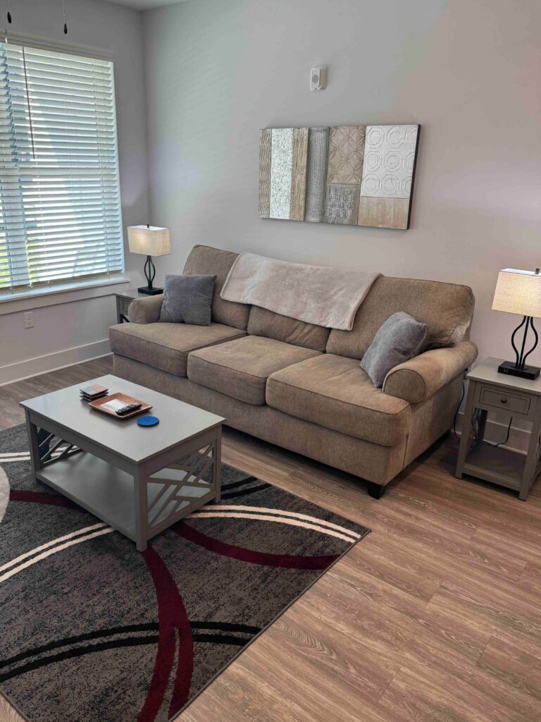 Modern living room featuring a comfortable sofa, coffee table, accent rug, end tables with lamps, large window, and contemporary neutral décor.