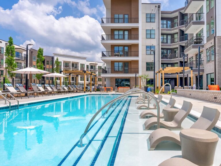 Resort-style outdoor swimming pool featuring in-water lounge chairs, expansive sundeck seating, shaded pergolas, and modern apartment buildings in the background.
