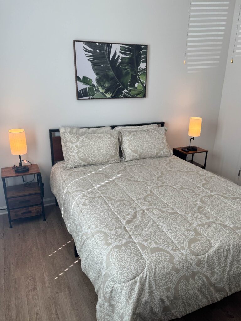 Furnished bedroom with patterned bedding, bedside lamps, wood-style flooring, and modern wall art at Exchange at Blair Stone.