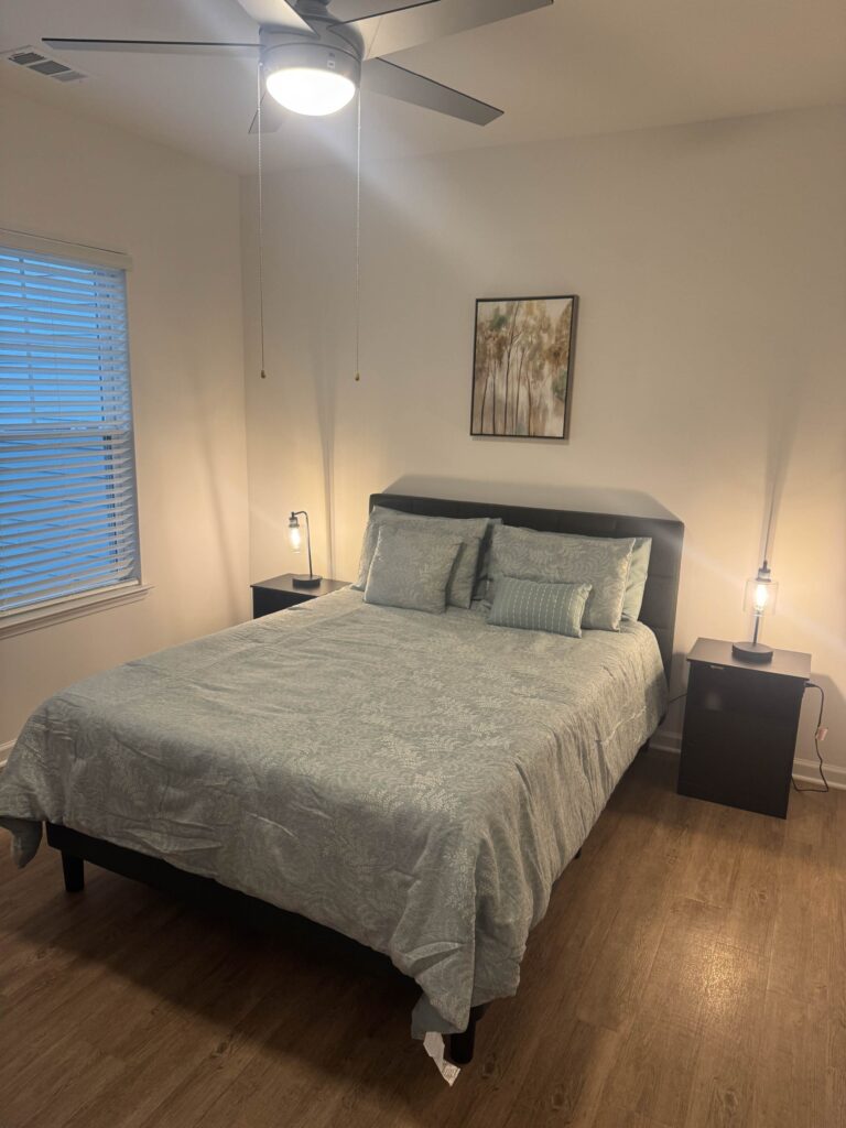 Modern furnished bedroom with queen bed, wood-style flooring, ceiling fan, and bedside lighting at Exchange at Blair Stone.