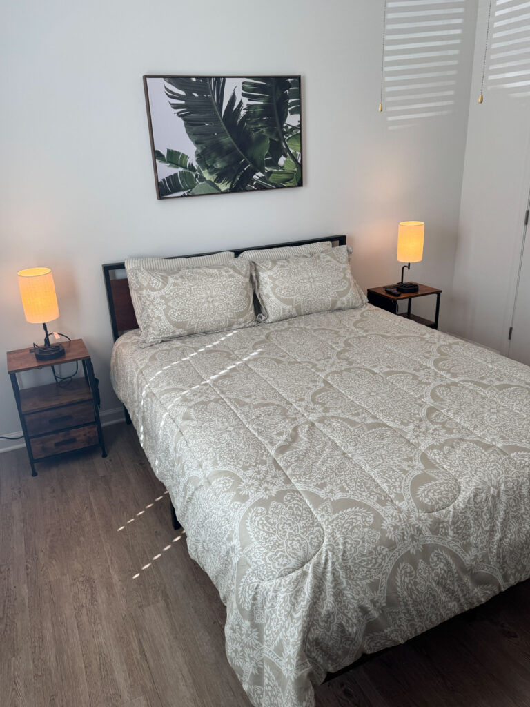 Modern furnished bedroom with queen bed, matching nightstands, warm lighting, and tropical wall art.