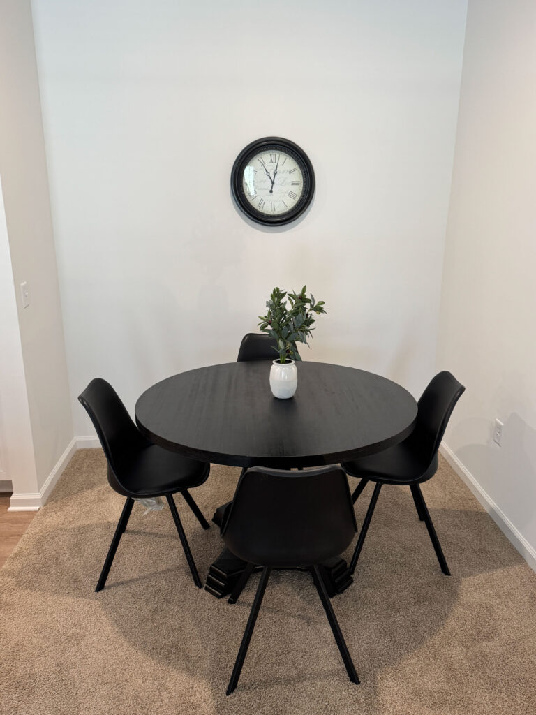 Modern dining area with round black table, four chairs, wall clock, and minimalist décor in a furnished apartment.