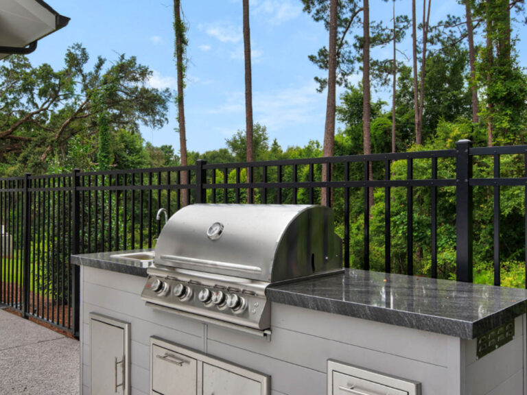 Outdoor grilling station with stainless steel barbecue grill, prep counter, and wooded backdrop behind a black metal fence.