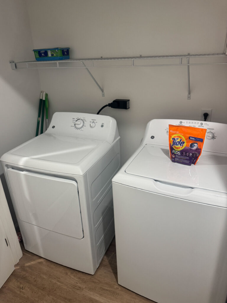 In unit laundry area with full size washer, dryer, shelving, and laundry supplies in a furnished apartment.