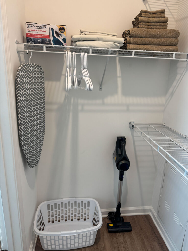 Walk in closet with storage shelving, folded linens, ironing board, vacuum, and laundry basket inside a furnished apartment.
