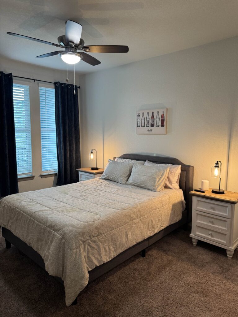 Modern furnished bedroom with a queen bed, bedside lighting, ceiling fan, and large windows with blackout curtains.