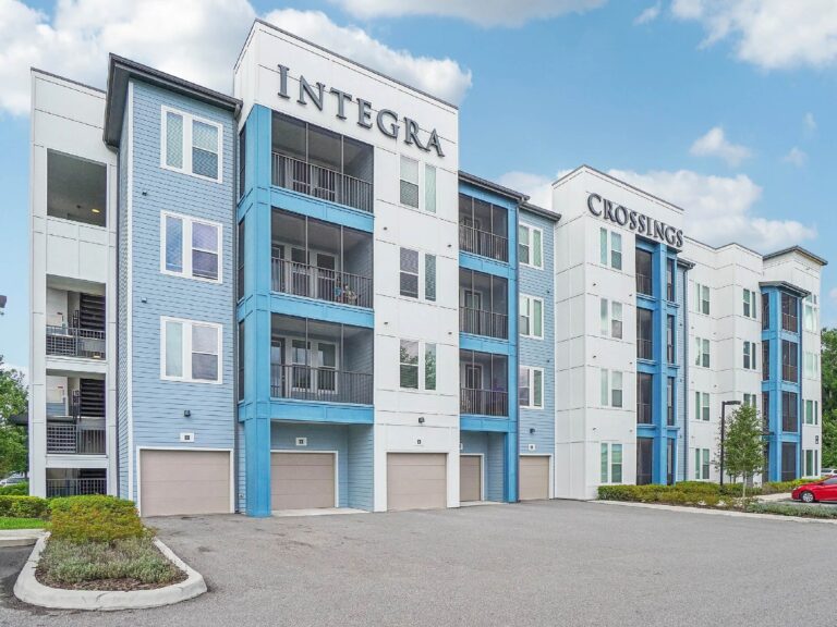 Modern apartment building exterior with blue and white accents, balconies, and attached garage spaces.
