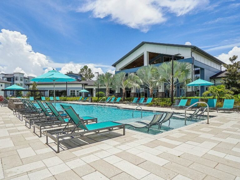 Resort-style swimming pool with lounge chairs, turquoise umbrellas, palm trees, and a modern clubhouse.