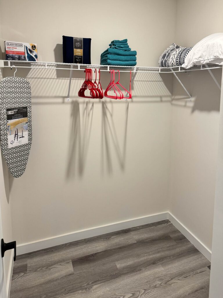 Walk-in storage closet with hanging space, shelving, ironing board, extra linens, towels, and guest convenience items.