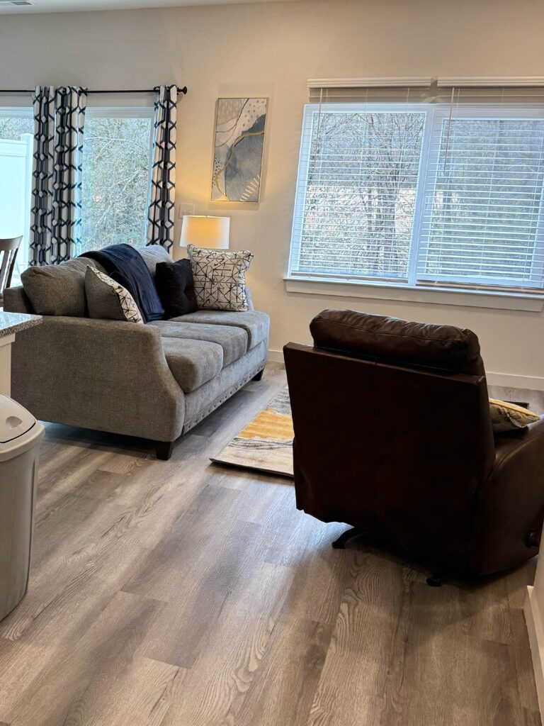 Bright living room featuring a comfortable gray sofa, leather recliner, large windows with natural light, luxury vinyl plank flooring, and contemporary décor.