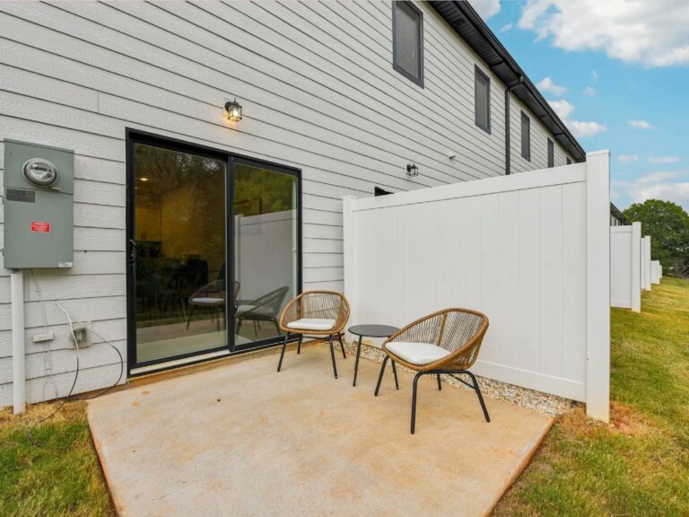 Private outdoor patio featuring two wicker-style chairs, a small table, sliding glass door access, and a privacy fence behind a modern townhome.