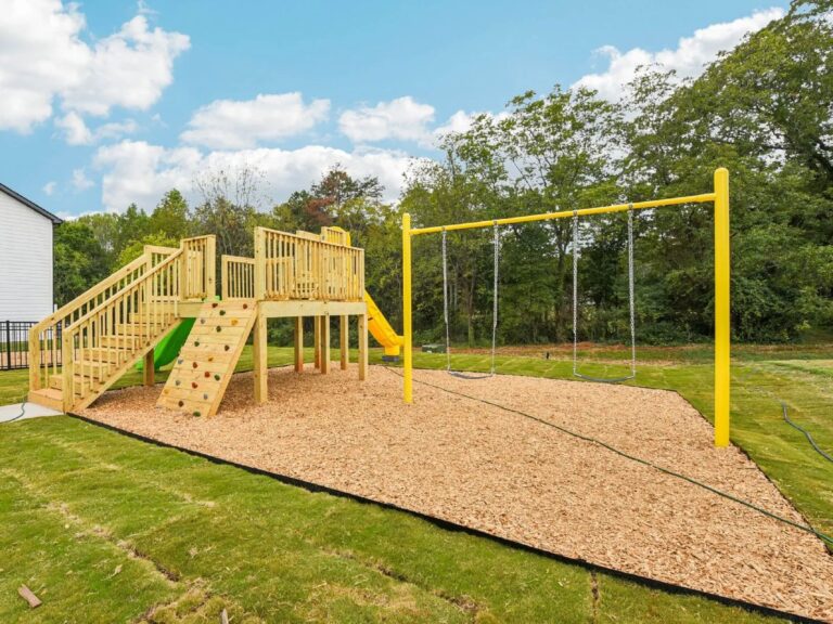 Community playground featuring swings, climbing wall, slides, and elevated play structures surrounded by open green space and mature trees.