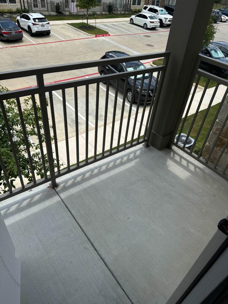 Private covered balcony at Lenox Grand Apartments in Austin, Texas featuring durable concrete flooring, metal railings, and views of the professionally maintained community grounds and parking areas.