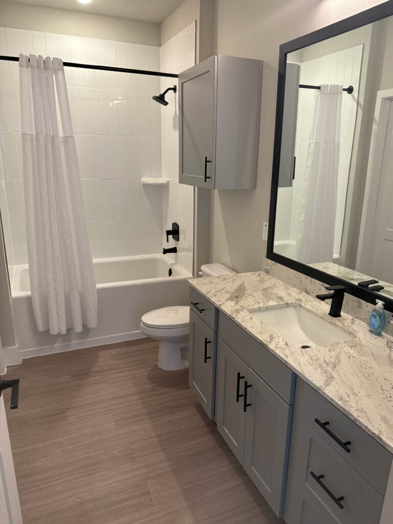 Modern full bathroom at Lenox Grand Apartments in Austin, Texas featuring a granite countertop vanity, black fixtures, white tiled tub and shower combination, large framed mirror, and luxury vinyl plank flooring.