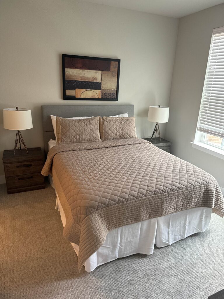 Fully furnished primary bedroom at Lenox Grand Apartments in Austin, Texas featuring a queen bed, upholstered headboard, matching nightstands, bedside lamps, soft carpeting, and contemporary wall art.