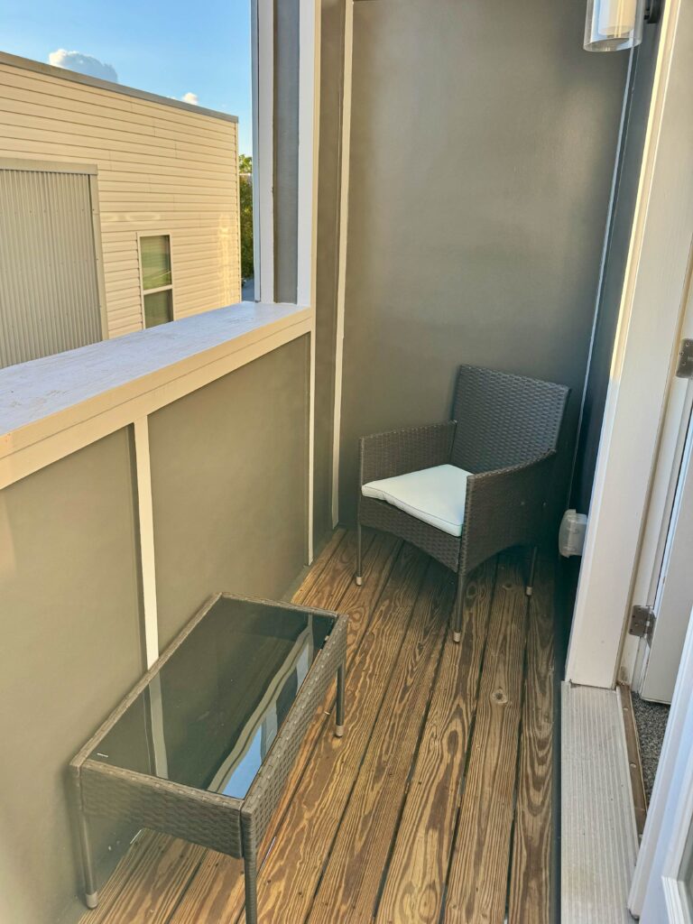 Private covered balcony featuring outdoor wicker seating, a glass-top table, wood plank flooring, and a peaceful elevated view of the surrounding community.