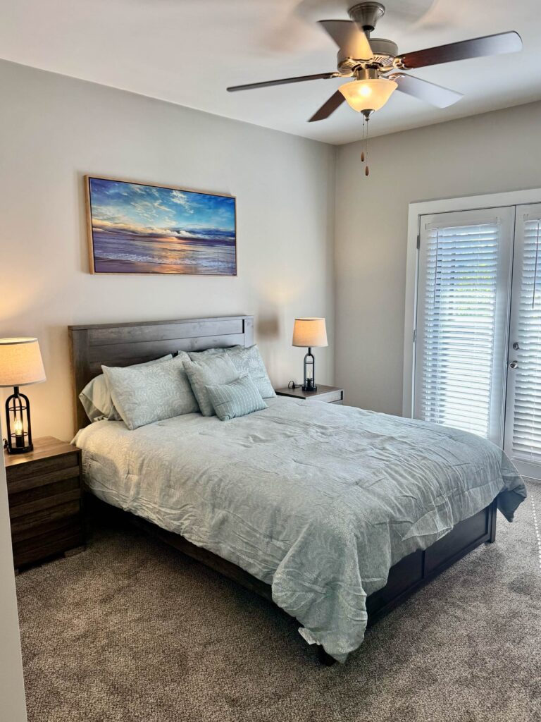 Spacious primary bedroom at Lofts at Rose Monte featuring a queen bed with coordinated bedding, matching nightstands with lamps, plush carpeting, ceiling fan, coastal artwork, and private access to a covered balcony through French doors.